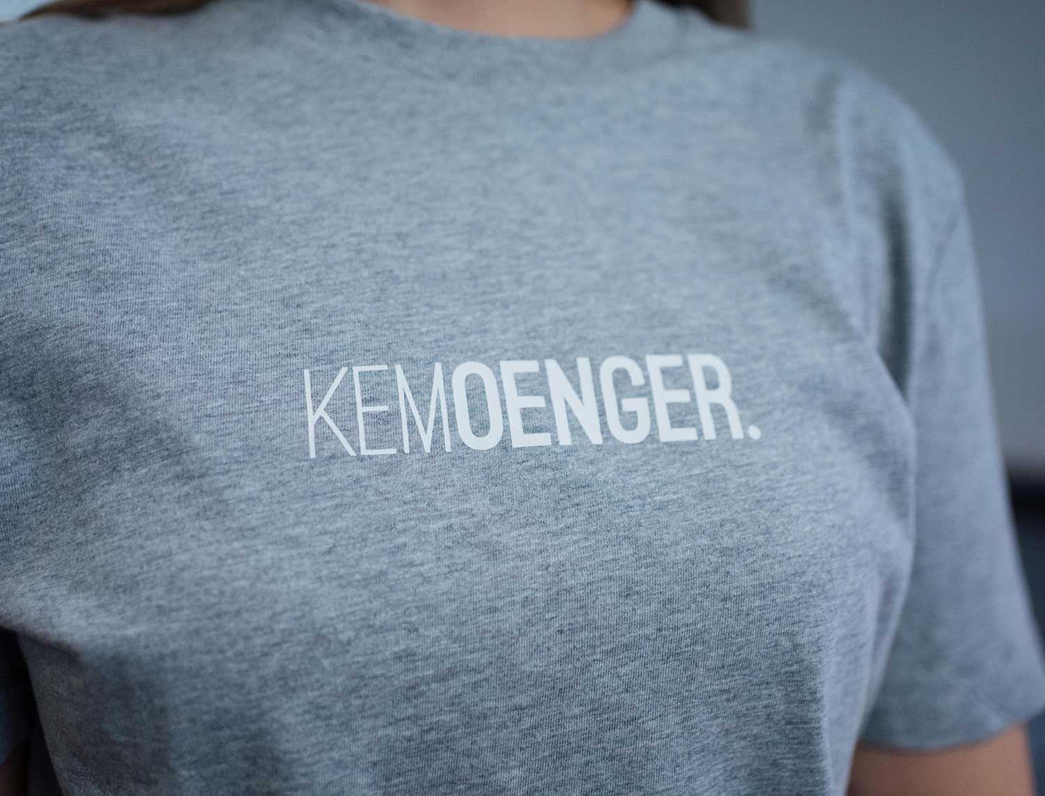 Close-up van een grijze T-shirt met opdruk KEMOENGER.