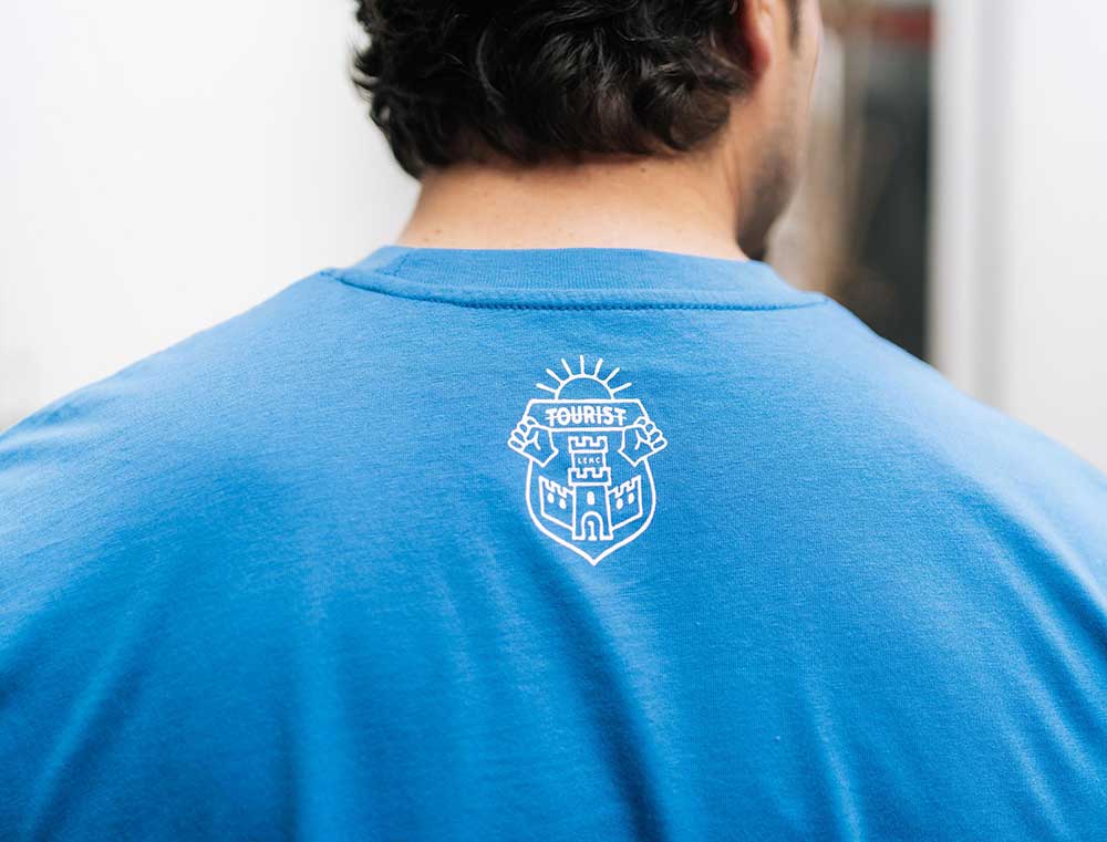 Achterkant van een blauwe T-shirt met het wapenschild van Tourist LeMC.