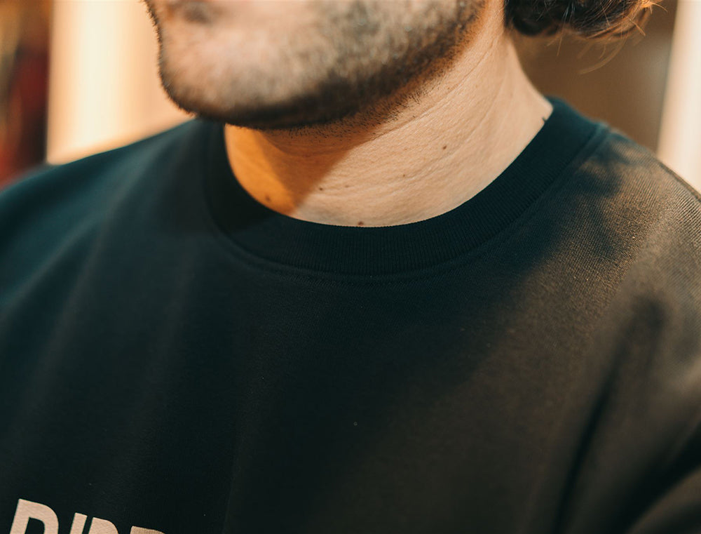 Close-up van pasvorm aan de nek van zwarte sweater met opdruk RIBBEDEBIE.
