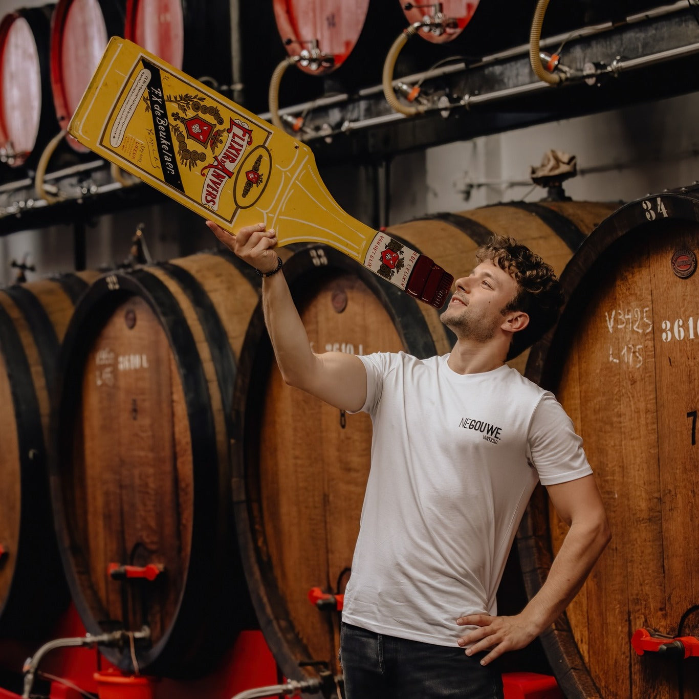 Man met een witte T-shirt met opdruk NEGOUWE. met in de hand een uitgesneden karton van Elixir d'Anvers en achter hem vaten bier