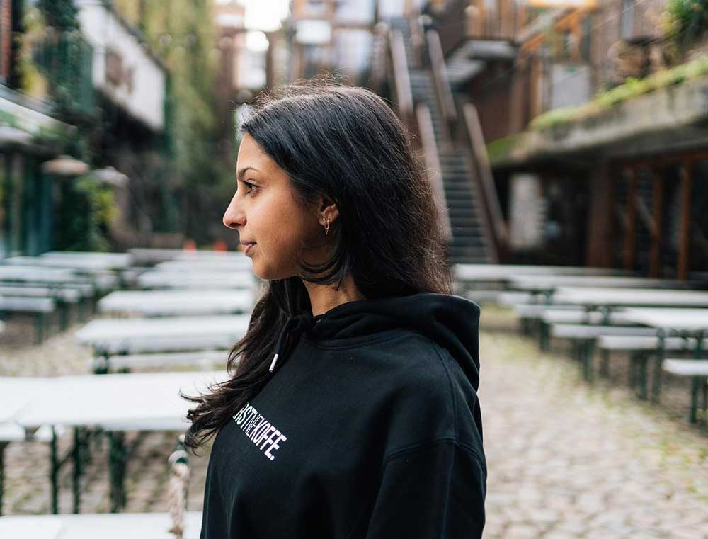 Zij-aanzicht van een vrouw met een zwarte hoodie met opdruk EERSTNEKOFFE.