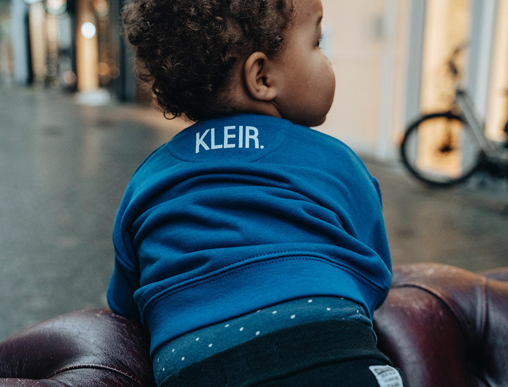 Baby met een blauwe baby-trui met opdruk KLEIR. in de nek