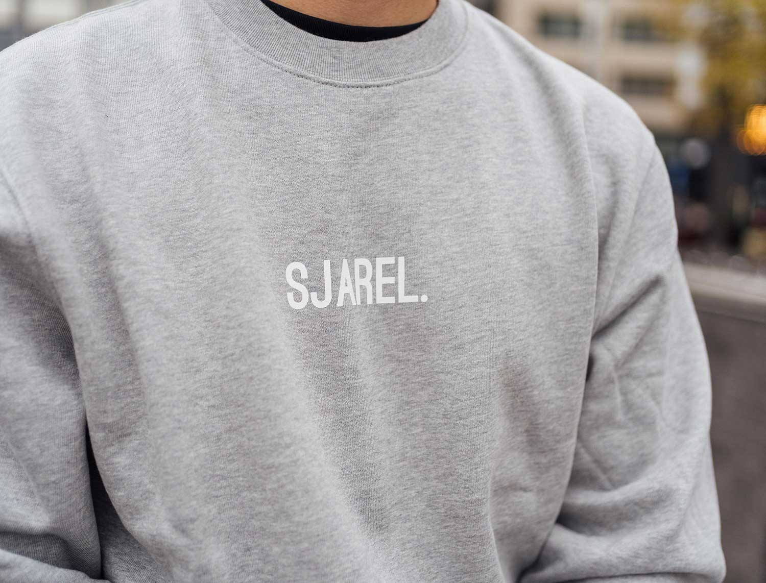 Close-up van een grijze sweater met opdruk SJAREL.