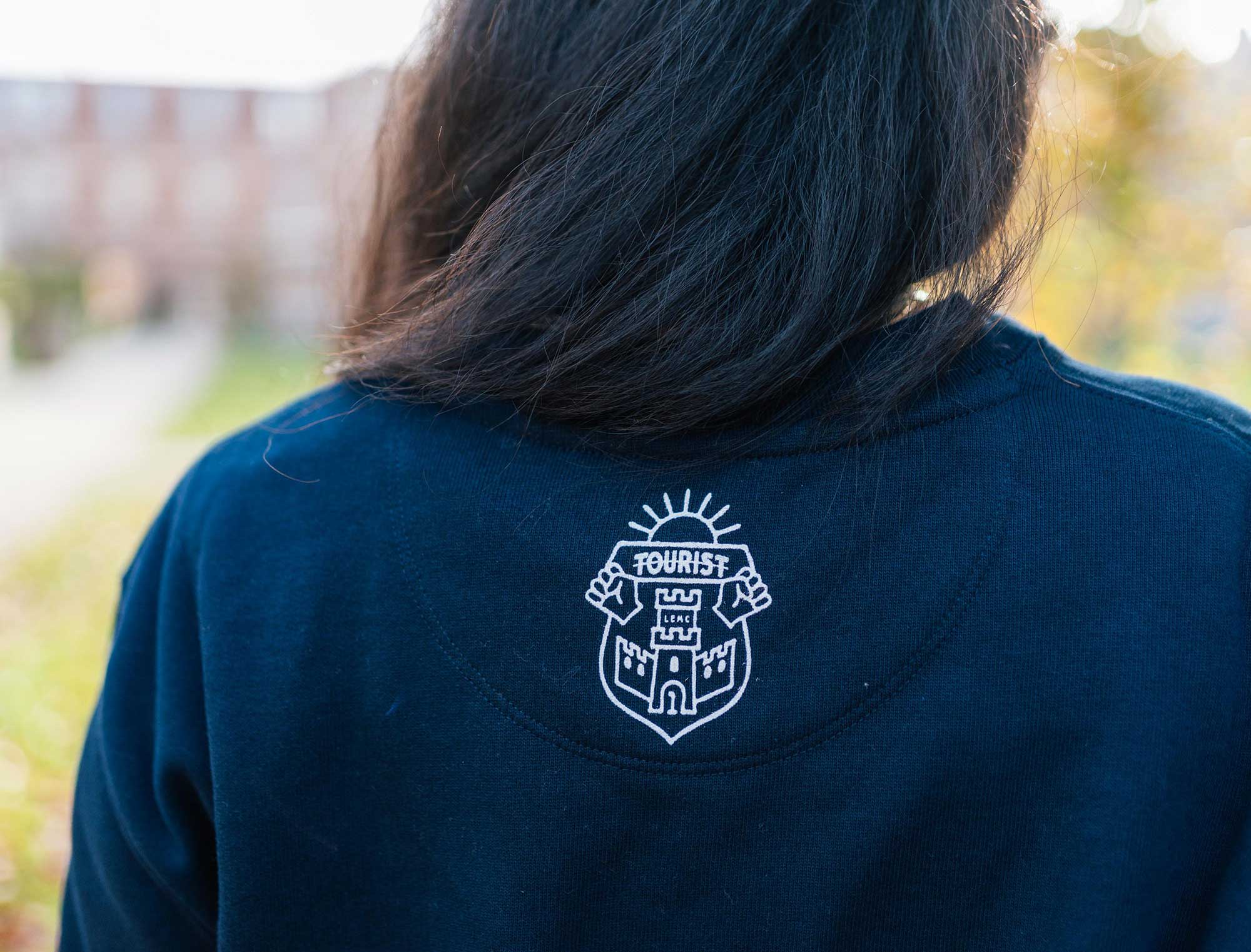 Achterkant van een blauwe sweater met het wapenschild van Tourist LeMC.