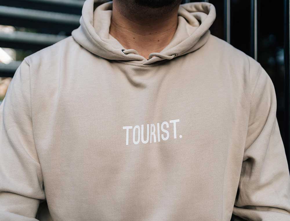 Close-up van een zandkleurige hoodie met opdruk TOURIST. vooraan.