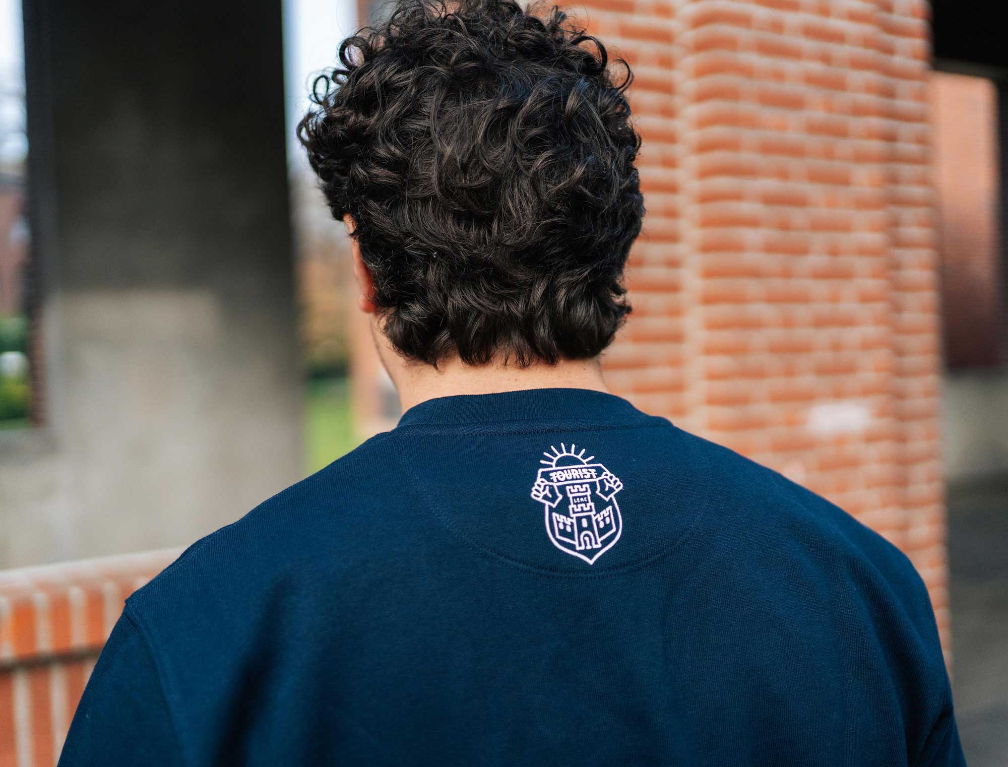 Achterkant van een blauwe sweater met het wapenschild van Tourist LeMC.