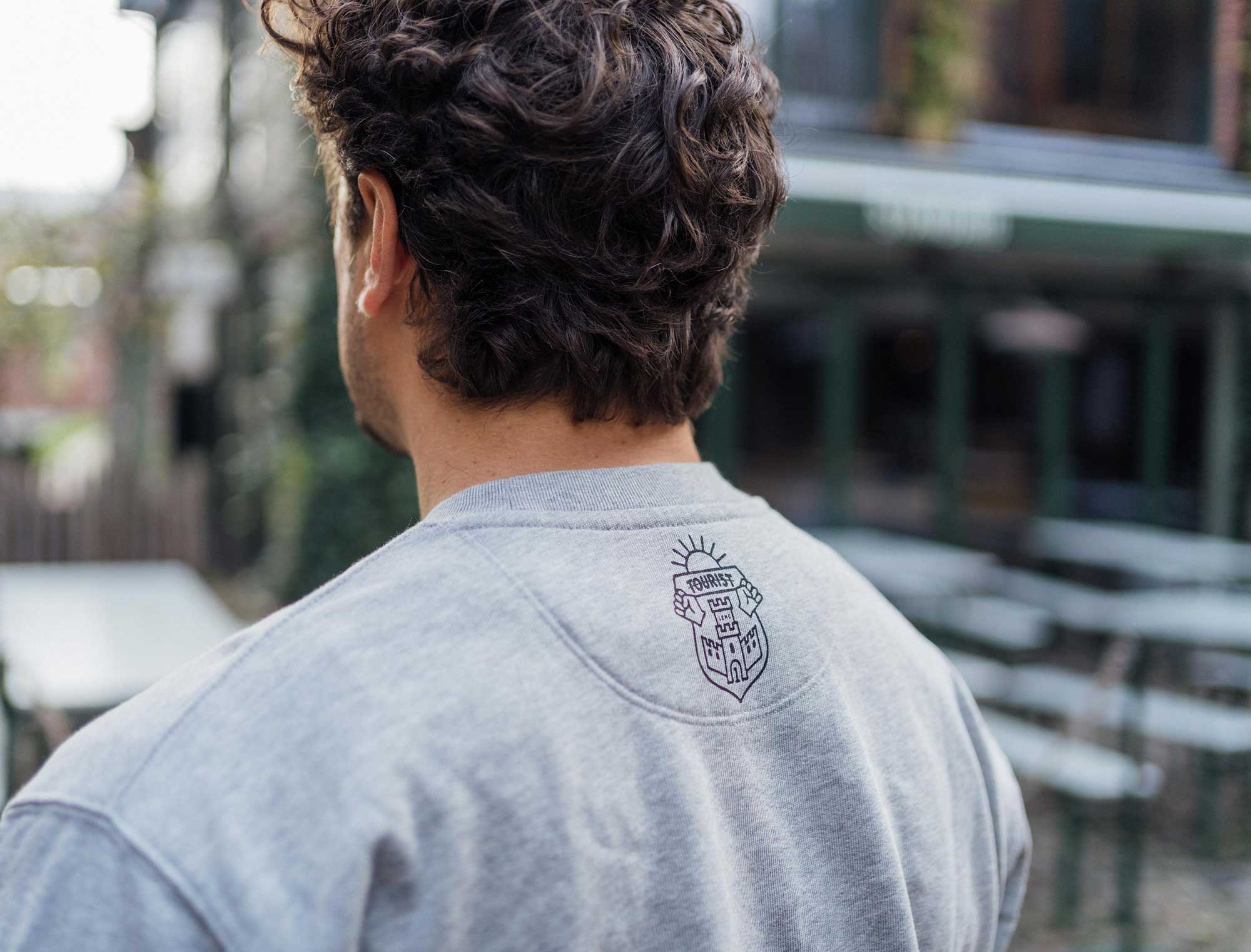 Achterkant van een grijze sweater met het wapenschild van Tourist LeMC.
